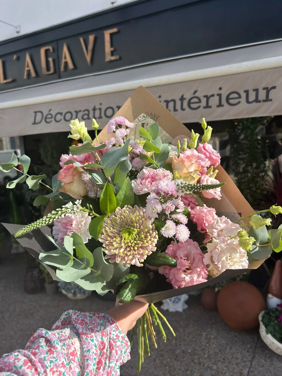 Bouquet pastel de lisianthus, roses pêche, œillets et eucalyptus emballé dans du papier kraft, tenu devant la façade noire de L’AGAVE à La Turballe