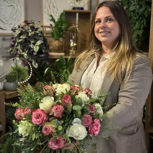 Une fleuriste de L’AGAVE sourit, un bouquet rond de roses roses et blanches à la main, dans la boutique