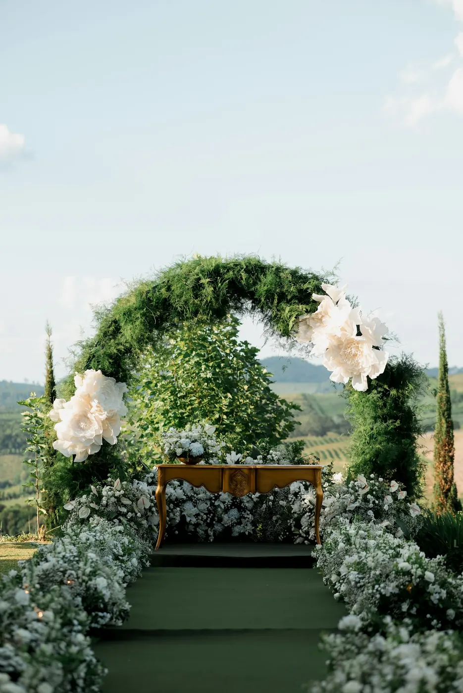Arche de cérémonie en feuillage et grandes fleurs blanches, au bout d’une allée bordée de fleurs blanches, en plein air