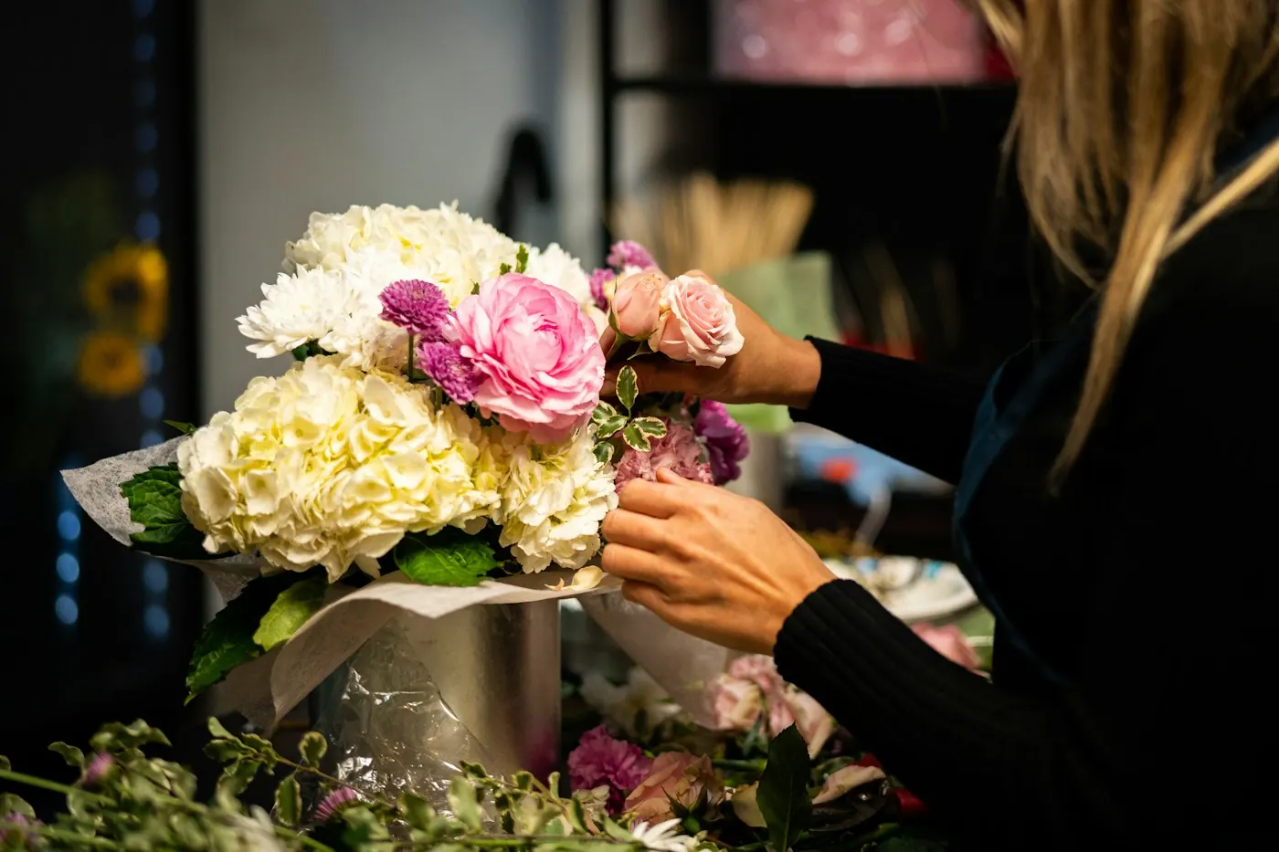 Deux mains arrangent un bouquet d’hortensias blancs et de roses roses, sur fond sombre
