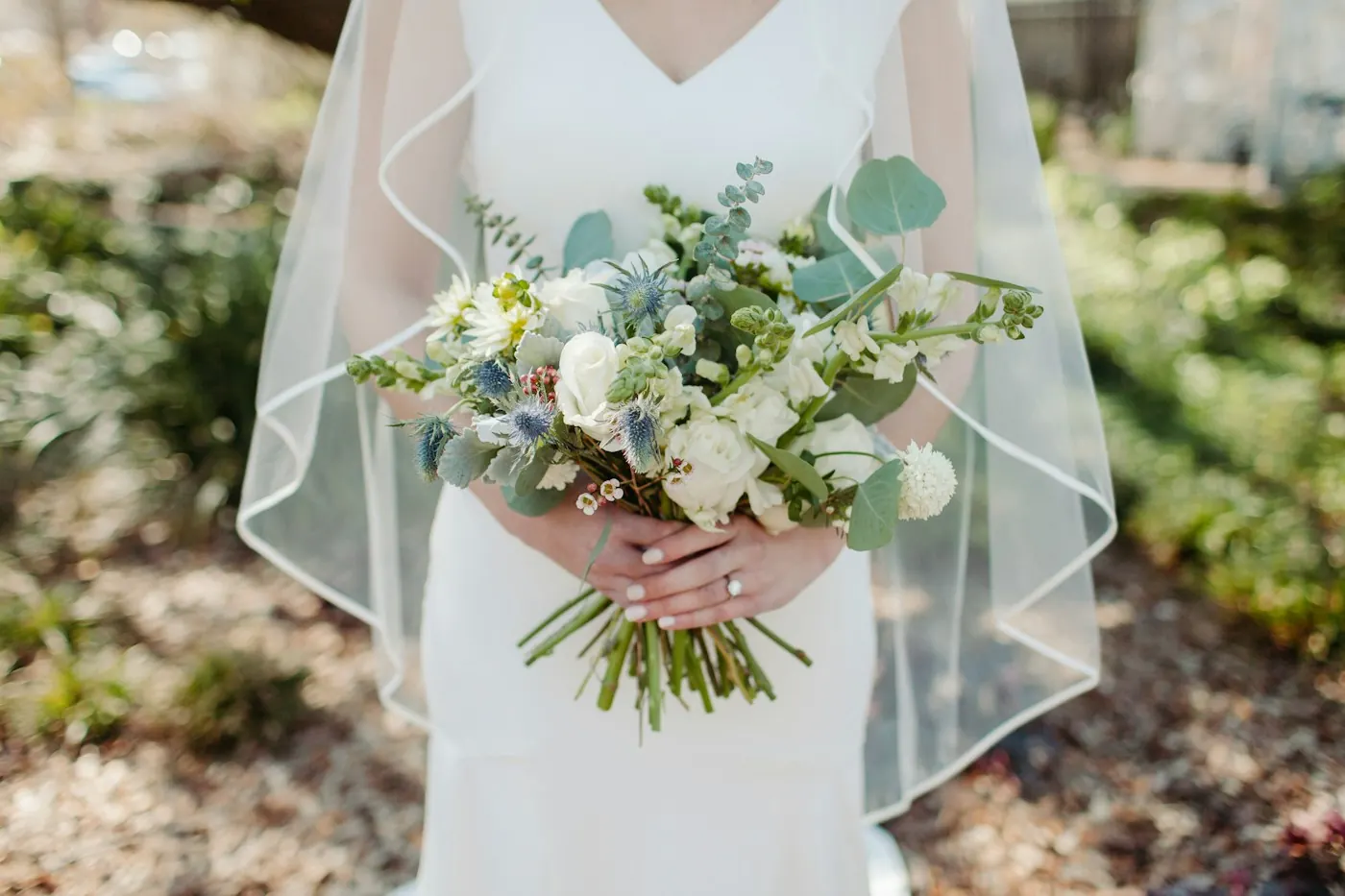 Mariée voilée tenant un bouquet de roses blanches, de chardons bleus et d’eucalyptus